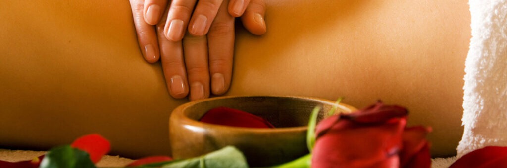 Hände bei der Rückenmassage, davor Holzschale mit roten Rosenblättern